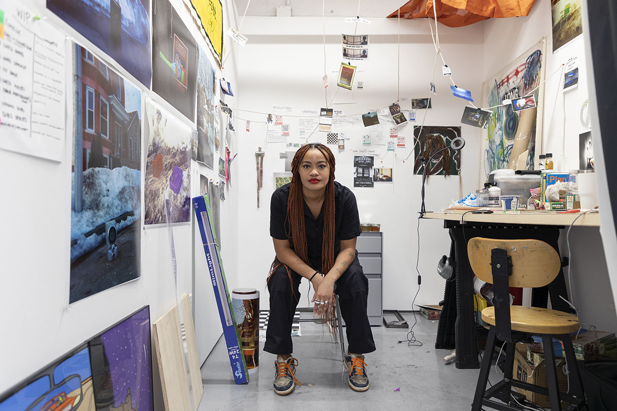 Photo of the artist seated in their studio. Photographs cover the walls and hand from the ceiling.