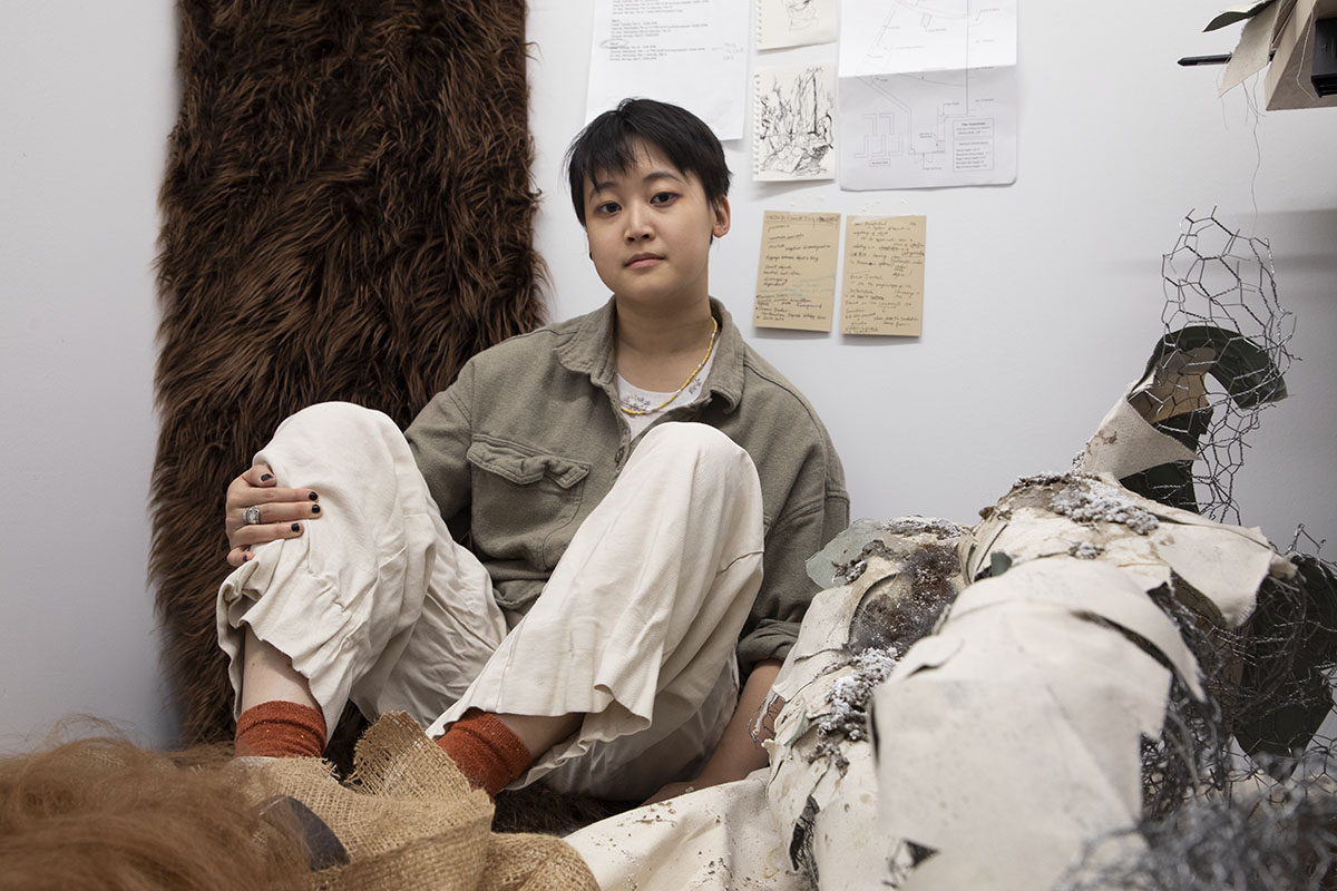 Photo of the artist in her studio with drawings on the wall behind her and fabric, chicken wire and other sculptural elements in the foreground.